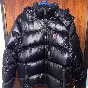 Svart dunjacka från Ralph Lauren - Säljer en svart, glansig dunjacka från Ralph Lauren med röd logga på bröstet. Jackan har huva, dragkedjor på fickorna och en quiltad design som ger en riktigt fet look. Perfekt för kalla dagar när du vill hålla stilen och värmen.köpte den för 5900kr för ett och ett halv år sedan har inte använt den någon gång,i så fall kanske 6 gånger.Pris går att diskutera. Till och med kostar jackan på Zalando 6600kr ny pris.