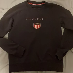Mörkblå sweatshirt från GANT med brodyr - Mörkblå sweatshirt från GANT med broderad logga och sköld med stjärnor och ränder på bröstet. Klassisk rund halsringning och långa ärmar. Mjuk och skön bomullsmix, perfekt för en stilren och sportig look. Priset är inte fast så det är bara att lägga ett bud!