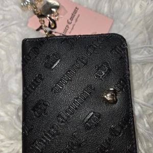 Juicy Couture svart plånbok med logga - Svart plånbok från Juicy Couture med präglad logga och kronor på utsidan, samt ett litet hjärtformat metallmärke. Insidan är beige med flera kortfack och fack för mynt, dekorerad med svart Juicy Couture-tryck. Guldig dragkedja och detaljer.