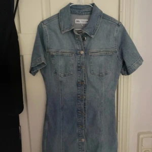 Kort jeansklänning från Zara - Snygg ljusblå jeansklänning från Zara med kort ärm, klassisk skjortkrage och knappar hela vägen framtill. Två bröstfickor ger en cool touch. Perfekt för dig som gillar denim och vill ha en avslappnad men trendig look.