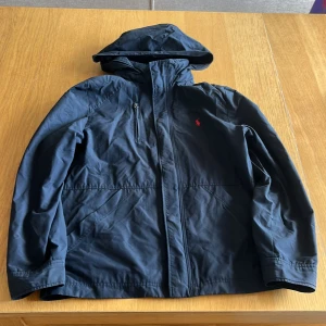 Mörkblå vindjacka från Polo Ralph Lauren - Snygg mörkblå vindjacka från Polo Ralph Lauren med huva och dragkedja framtill. Storlek L i kid size och XS i men size