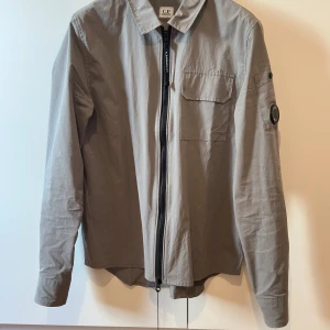 Grå overshirt från C.P. Company - Snygg grå overshirt från C.P. Company med klassisk krage, bröstficka med lock och ikonisk linsdetalj på ärmen. Jackan har dragkedja framtill och är tillverkad i bomull. Perfekt för dig som gillar stilrena och funktionella plagg.