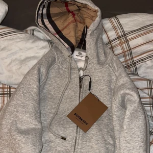 Grå hoodie från Burberry med dragkedja - Snygg grå hoodie från Burberry med klassiskt rutigt foder i huvan. Tröjan har dragkedja framtill, snörning vid huvan och ribbade muddar. Perfekt för dig som gillar stilrena märkesplagg med exklusiv känsla.