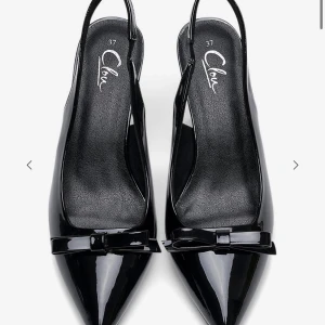 Svarta lackpumps med rosett från Clou - Snygga svarta pumps i lack från clou med spetsig tå och elegant rosettdetalj framtill. Skorna har öppen häl med rem och en smal klack som ger en stilren siluett. Storlek 39. Aldrig använda, säljer då det ör för sent att lämna tbx dom 