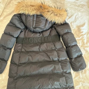 Svart Moncler dunjacka med pälskrage - Säljer en svart lång Moncler dunjacka med snygg pälskantad huva och markerat midjeband med logga. Jackan har dragkedjor framtill och på fickorna, quiltad design och en riktigt lyxig vibe. Perfekt för kalla dagar när du vill vara både varm och stilren. Jackan är i EU storlek M, kinesisk storlek XXL. 