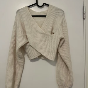 Beige omlott stickad croppad tröja - Superfin beige stickad tröja med omlott-design och croppad passform. Tröjan har bred v-ringning och långa ärmar med ribbad struktur. Perfekt för lager-på-lager och ger en chill men ändå trendig vibe.