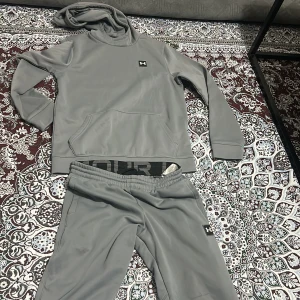 Grå hoodie från Under Armour - Säljer en stilren grå hoodie från Under Armour med huva och magficka. Tröjan har lång ärm, diskret logga på bröstet och stor Under Armour-text baktill. Perfekt för träning eller chill. Materialet är mjukt och stretchigt polyester.