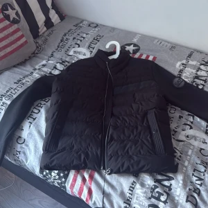 Svart pufferjacka Emporio Armani EA7 - Snygg svart pufferjacka från Emporio Armani EA7 med quiltad front, släta ärmar och dragkedja. Jackan har diskret logga på ärmen och detaljerad Emporio Armani-text på bröstet. Perfekt för dig som vill ha en stilren och modern look. Den är helt ny och oanvänd med tagg på. Pris kan diskuteras