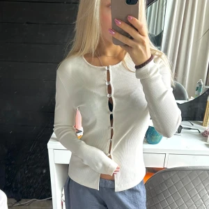 Vit ribbad kofta med hjärtknappar - Supersöt vit ribbad kofta med långa ärmar och unika silvriga hjärtformade knappar framtill. Sitter snyggt och har en lätt croppad passform. Perfekt att styla med jeans eller kjol för en trendig look.