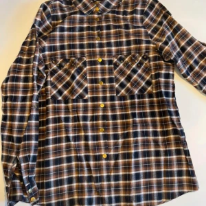 Rutig skjorta med nitar från Gina Tricot - Rutig skjorta i bruna nyanser från Gina Tricot. Skjortan har långa ärmar, två bröstfickor med coola nitar och guldiga tryckknappar framtill. Perfekt för dig som gillar en avslappnad men edgy stil.