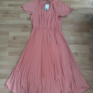Ny. Plisserad korallrosa långklänning H&M - Superfin långklänning med fider från H&M i en härlig korallrosa nyans. Klänningen har plisserad kjol, korta ärmar och knytning vid halsen. Den är luftig och har en markerad midja som ger en snygg siluett. Perfekt för dig som vill sticka ut med en färgstark look. Aldrig använd.