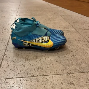 Nike Mercurial blå fotbollsskor - Säljer ett par Nike Mercurial fotbollsskor i blått med gula detaljer och grafiskt mönster. Skorna har elastisk snörning, låg profil och plös med kardborre. Sulan är blå med gula dobbar, perfekt för gräsplan. Materialet är syntet och skon har en sportig vibe.