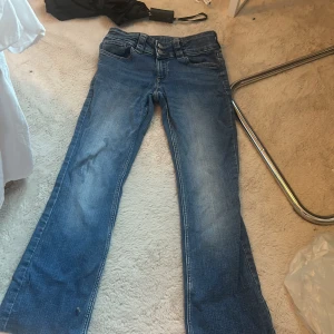 Blå bootcut jeans med dubbla knappar - Snygga blå jeans med bootcut-modell och dubbla silverfärgade knappar i midjan. Jeansen har klassisk femficksdesign och markerade sömmar. Perfekta för dig som gillar en retrovibe och vill ha ett par jeans med lite utsvängda ben.