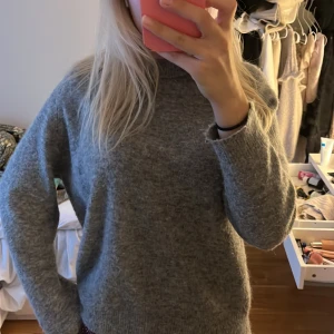 Grå stickad tröja - Mysig grå stickad tröja med hög hals och lång ärm. Perfekt för kyliga dagar och enkel att matcha med jeans eller kjol. Tröjan har en loose fit och är tillverkad i ett mjukt material som känns skönt mot huden.