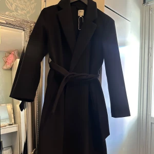 Svart kappa från ONLY med bälte - Stilren svart kappa från ONLY med klassisk krage och knytskärp i midjan. Lång modell med raka ärmar och enkel design som ger en clean look. Perfekt för dig som gillar minimalistisk stil och vill ha en jacka som funkar till allt.