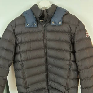 Mörkbrun Colmar dunjacka med blå krage - Säljer en snygg mörkbrun dunjacka från Colmar med blå krage och diskret logga på ärmen. Jackan har horisontella quiltade sektioner, två fickor med dragkedja och hel dragkedja framtill. Perfekt för kalla dagar och har en modern, sportig look.