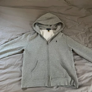 Grå hoodie från Polo Ralph Lauren XL - Grå hoodie från Polo Ralph Lauren i storlek XL. Tröjan har dragkedja, huva och två fickor framtill. Insidan är mjuk och fleecefodrad för extra komfort. Klassisk broderad logga på bröstet och enkel, stilren design. Liten skada vid dragkjedjan.