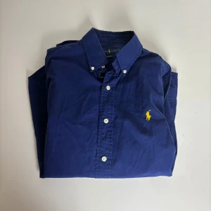 Blå skjorta från Ralph Lauren S - Klassisk marinblå skjorta från Ralph Lauren i slim fit med knappar framtill och button down-krage. Skjortan har det ikoniska gula broderade Polo-loggan på bröstet och är tillverkad i mjuk bomull. Perfekt för en clean och stilren look.