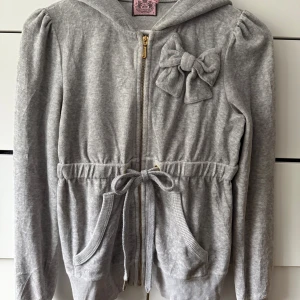 Vintage Juicy Couture Hoodie - Superfin grå hoodie från Juicy Couture med dragkedja, huva och en söt rosett på bröstet. Tröjan har knyt i midjan, ribbade muddar och två fickor framtill.