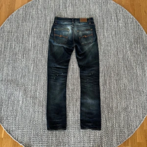 Nudie Jeans blå slitna jeans 29/32 - Säljer ett par blå jeans från Nudie Jeans med snygga slitningar och hål på benen. Klassisk femficksmodell med orangea sömmar och läderpatch bak. Jeansen har en rak passform och är tillverkade i bomull. Perfekta för dig som gillar en avslappnad och trendig look.