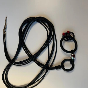 Svart band med ringar och röda detaljer - Unikt svart halsband med flera stora och små ringar i gummi, dekorerat med två röda kulor och en silverfärgad metallpärla. Långa svarta snören med metalländar ger en cool och modern vibe. Perfekt för dig som gillar statement-smycken.