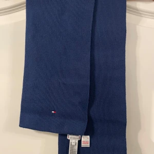 Blå halsduk från Tommy Hilfiger - En stilren blå halsduk från Tommy Hilfiger med diskret logga broderad i ena hörnet. Halsduken är ribbstickad och tillverkad i 100% bomull, vilket gör den både mjuk och bekväm. Perfekt accessoar för att lyfta din outfit under kyliga dagar. Helt oanvänd!