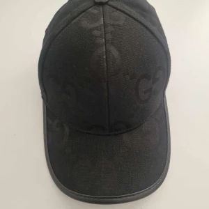 Svart keps med GG-mönster - Säljer en svart keps från med diskret GG-monogram över hela kepsen. Kepsen har böjd skärm, justerbart spänne bak i skinn och är tillverkad i ett slitstarkt bomullsmaterial. Snygg och stilren accessoar för dig som gillar lyxiga detaljer.