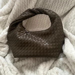 Brun flätad axelväska i skinn - Snygg brun axelväska med flätad design och dragkedja. Väskan har en mjuk, böjd form och är tillverkad i skinn. Perfekt för dig som gillar stilrena accessoarer med en twist.