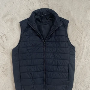 Mörkblå quiltad dunväst från Zara - Stilren mörkblå dunväst från Zara med quiltad design och hög krage. Västen har dragkedja framtill och är perfekt för lager-på-lager. Enkel och clean look som funkar till många outfits.