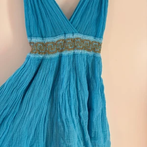 Turkos kortklänning med guldmönster - Superfin turkos kortklänning i boho-stil med omlott-v-ringning och bred midjedetalj i guldigt, grekiskt mönster. Klänningen är sydd i ett lätt, krinklat tyg som ger en luftig känsla. Perfekt för sommarens varma dagar och har en riktigt snygg passform. 