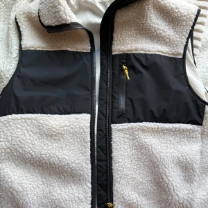 Vit och svart fleeceväst från H&M - Snygg väst från H&M i vit teddyfleece med svarta paneler och detaljer. Västen har dragkedja framtill, bröstficka med gul dragkedja och hög krage. Perfekt för lager-på-lager och chill stil under kyliga dagar.