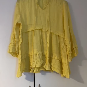 Gul volangblus med knytband - Säljer en söt gul blus med volanger och knytband framtill. Blusen har långa ärmar med volangdetaljer och en lös, flowy passform. Perfekt för dig som gillar färg och en bohemisk vibe.
