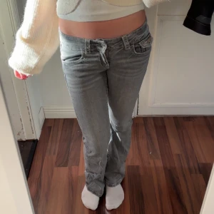 Grå bootcut jeans med låg midja - Säljer ett par grå jeans med låg midja och bootcut-ben. Jeansen har snygga detaljer som fickor med lock och knapp bak samt klassisk femficksdesign. Perfekta för dig som gillar en avslappnad men trendig look.