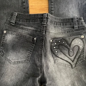 Svarta Justice jeans med hjärtdetalj - Snygga svarta jeans från Justice med slitningseffekt och coolt hjärta i svart brodyr och nitar på bakfickan. Klassisk femficksmodell med dragkedja och bälteshällor. Perfekta för dig som vill sticka ut lite extra.