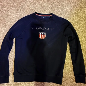 Mörkblå sweatshirt från GANT - Säljer en mörkblå sweatshirt från GANT med broderad logga och sköld på bröstet. Tröjan har rund halsringning, långa ärmar och ribbade muddar. Klassisk och stilren design med röd och vit detalj på insidan av kragen. Ny pris 400kr mitt pris 250kr