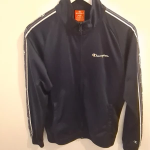 Mörkblå track jacket från Champion - Snygg mörkblå track jacket från Champion med vit logga broderad på bröstet och loggor längs ärmarna. Jackan har dragkedja framtill, ståkrage och två fickor. Tillverkad i glansig polyester för en sportig vibe.
