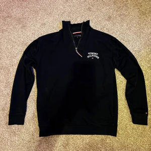 Svart Tommy Hilfiger half zip tröja - Svart långärmad tröja från Tommy Hilfiger med broderad logga på bröstet och liten flagga på ärmen. Tröjan har en snygg half zip-dragkedja vid halsen och ribbade muddar. Perfekt för dig som gillar klassisk och stilren streetwear. Ny pris 1599kr mitt pris 599