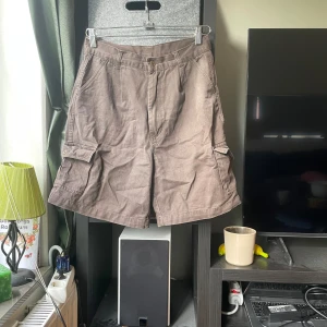 Bruna cargo shorts från Folk Collection - Säljer ett par bruna cargo shorts från Folk Collection i storlek S. De har flera fickor med lock på sidorna och bak, klassisk knapp och dragkedja framtill samt avslappnad passform. Perfekta för chill dagar och har en enkel, stilren look.