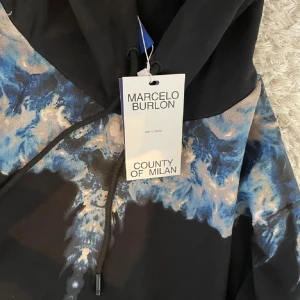 Svart hoodie från Marcelo Burlon - Säljer en svart hoodie från Marcelo Burlon County of Milan med coolt blått och beige mönster över axlar och bröst. Tröjan har huva med snörning och en stor magficka. Perfekt för dig som vill sticka ut med en unik design.