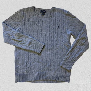 Grå stickad tröja från Polo Ralph Lauren - Snygg grå stickad tröja från Polo Ralph Lauren i storlek Barn xl, så den sitter som en S. Tröjan har kabelstickat mönster, rund hals och långärmade ribbade muddar. Klassisk Polo-logga broderad på bröstet. Perfekt för dig som gillar stilrena och tidlösa plagg.