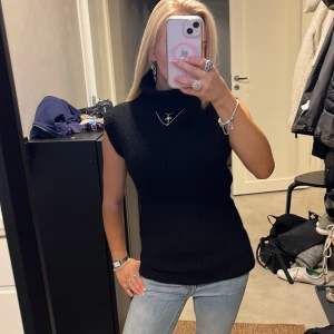 Svart stickad väst med polokrage - Säljer en svart stickad väst med polokrage och ribbad struktur. Västen är ärmlös och har en clean, slim passform som sitter snyggt på kroppen. Perfekt att styla över en skjorta eller t-shirt för en trendig look.