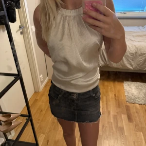 Vit ärmlös blus med halterneck - Säljer en vit blus med halterneck och lätt glansig yta. Toppen är ärmlös och har en lös passform som kan stylas både instoppad och utanför byxan. Materialet känns svalt och luftigt, perfekt för varmare dagar.