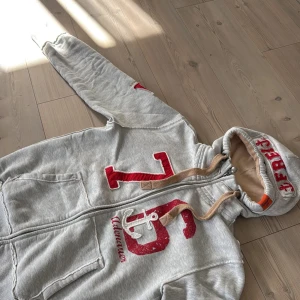 Sällsynt unik hoodie från Adenauer & Co - Säljer en grå hoodie från Adenauer & Co med stor röd 76-print och ankare på bröstet. Tröjan har huva med snörning, dragkedja framtill och två fickor. Muddar vid ärmslut och nederkant ger en skön passform. Går knappt att hitta och nypris på crika 2 tusen kr . Denna tröja är sällsynt, har inte sett något från detta märke som är relativt exklusivt här på plick!