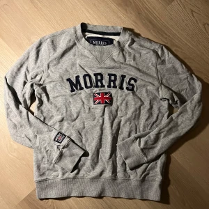 Grå sweatshirt från Morris, small - Grå sweatshirt från Morris med broderad logga och brittisk flagga på bröstet. Tröjan har rund hals, ribbade muddar och en patch på ärmen. Perfekt för en chill och stilren look.