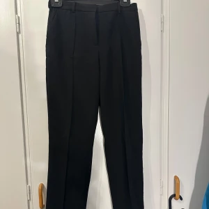 Svarta kostymbyxor från H&M - Klassiska svarta kostymbyxor från H&M med raka ben, pressveck och hällor för bälte. Byxorna har dragkedja och knappstängning framtill samt diskreta bakfickor. Perfekta för en clean och stilren look.