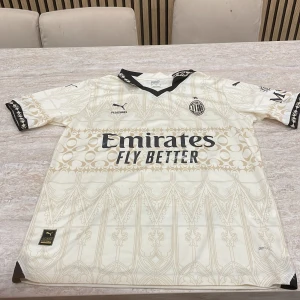 AC Milan x Puma fotbollströja S - AC Milan fotbollströja från Puma i ljusbeige med guldiga detaljer och grafiskt mönster. Svart V-ringad krage och svarta detaljer på ärmsluten. Materialet är lätt och andas, perfekt för match eller träning. Stora tryck med Emirates och klubbmärke på bröstet.