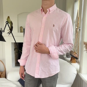 Rosa randig skjorta från Polo Ralph Lauren - Snygg rosa skjorta från Polo Ralph Lauren med vita vertikala ränder och broderad logga på bröstet. Slim fit-modell med klassisk krage och knäppning framtill. Tillverkad i mjuk bomull och har långa ärmar med manschett.