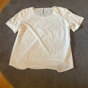 Vit blus med spetsärmar - Säljer en söt vit blus med korta ärmar i spets och rund halsringning. Blusen har en rak passform och en liten knapp i nacken. 