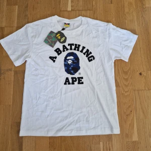 Vit BAPE t-shirt med blå camo print - Säljer en clean vit t-shirt från BAPE i storlek L. Framsidan har det klassiska 'A Bathing Ape'-trycket med blå camo-mönstrad ape-logo. T-shirten har rund hals, korta ärmar och är gjord i mjuk bomull. Taggar och BAPE-märke på nederkanten ingår. Storlek L, lite liten I storlek !