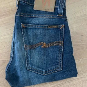 Blå jeans från Nudie Jeans - Snygga blå jeans från Nudie Jeans med klassisk femficksdesign och orangea sömmar på bakfickorna. Jeansen har en rak passform och är tillverkade i slitstarkt denimtyg av bomull. Perfekta för dig som gillar en tidlös och stilren look. Riktigt snygga jeans, storlek 26/32 som ni ser på bilden. De är som nya, frakt sker samma dag som köp! Va inte rädd att ta kontakt!